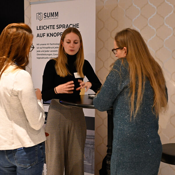 Drei Frauen stehen um einen Tisch herum. Die Frau in der Mitte erklärt etwas, die anderen beiden hören ihr zu. Im Hintergrund ist das Logo von SUMM AI auf einer Stellwand zu sehen, darunter Marketingtext.
