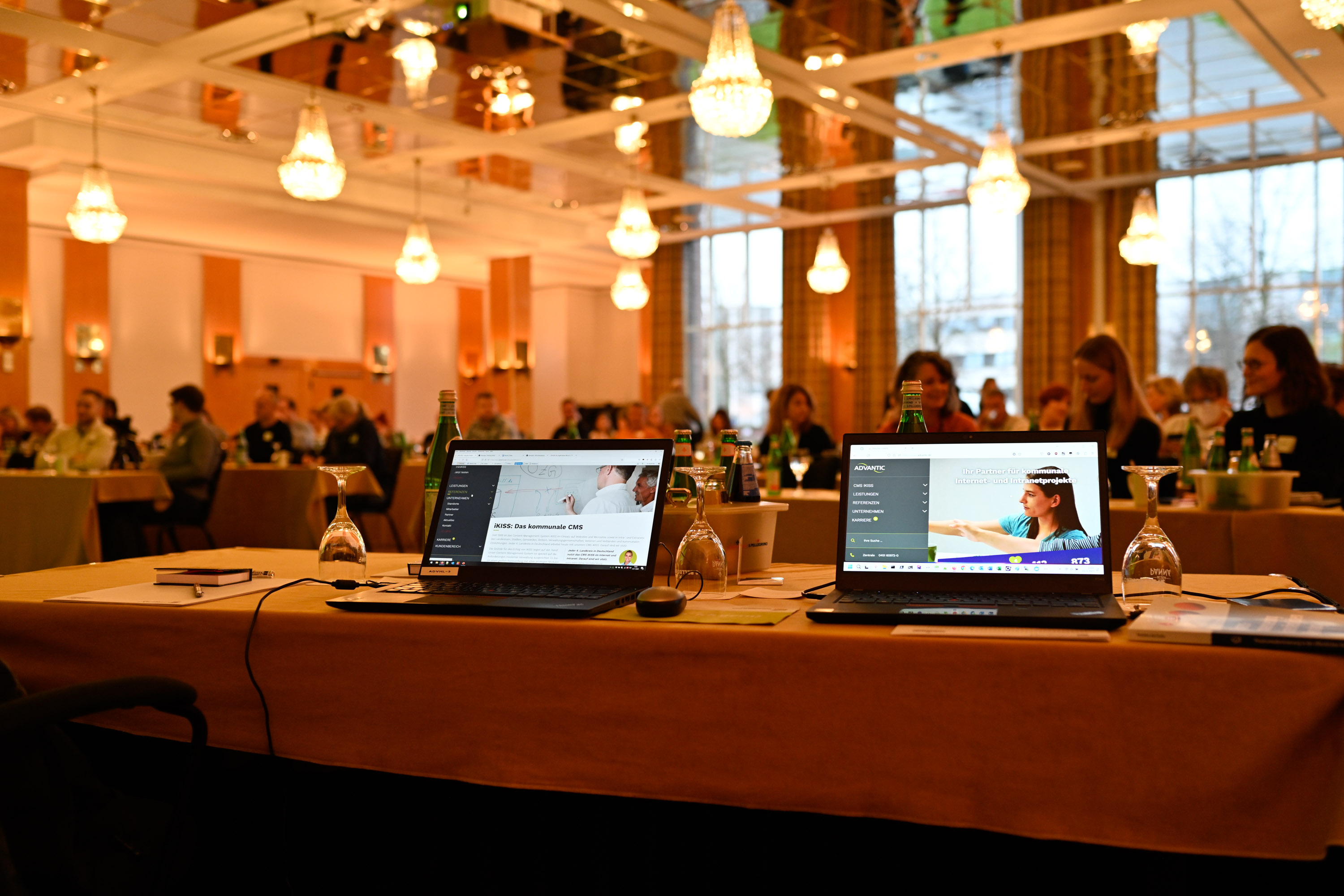 Blick in einen Konferenzraum, locker gefüllt mit Menschen. Im Vordergrund stehen zwei aufgeklappte Laptops mit der geöffneten Website der Advantic GmbH auf den Displays.
