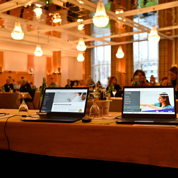 Blick in einen Konferenzraum, locker gefüllt mit Menschen. Im Vordergrund stehen zwei aufgeklappte Laptops mit der geöffneten Website der Advantic GmbH auf den Displays.